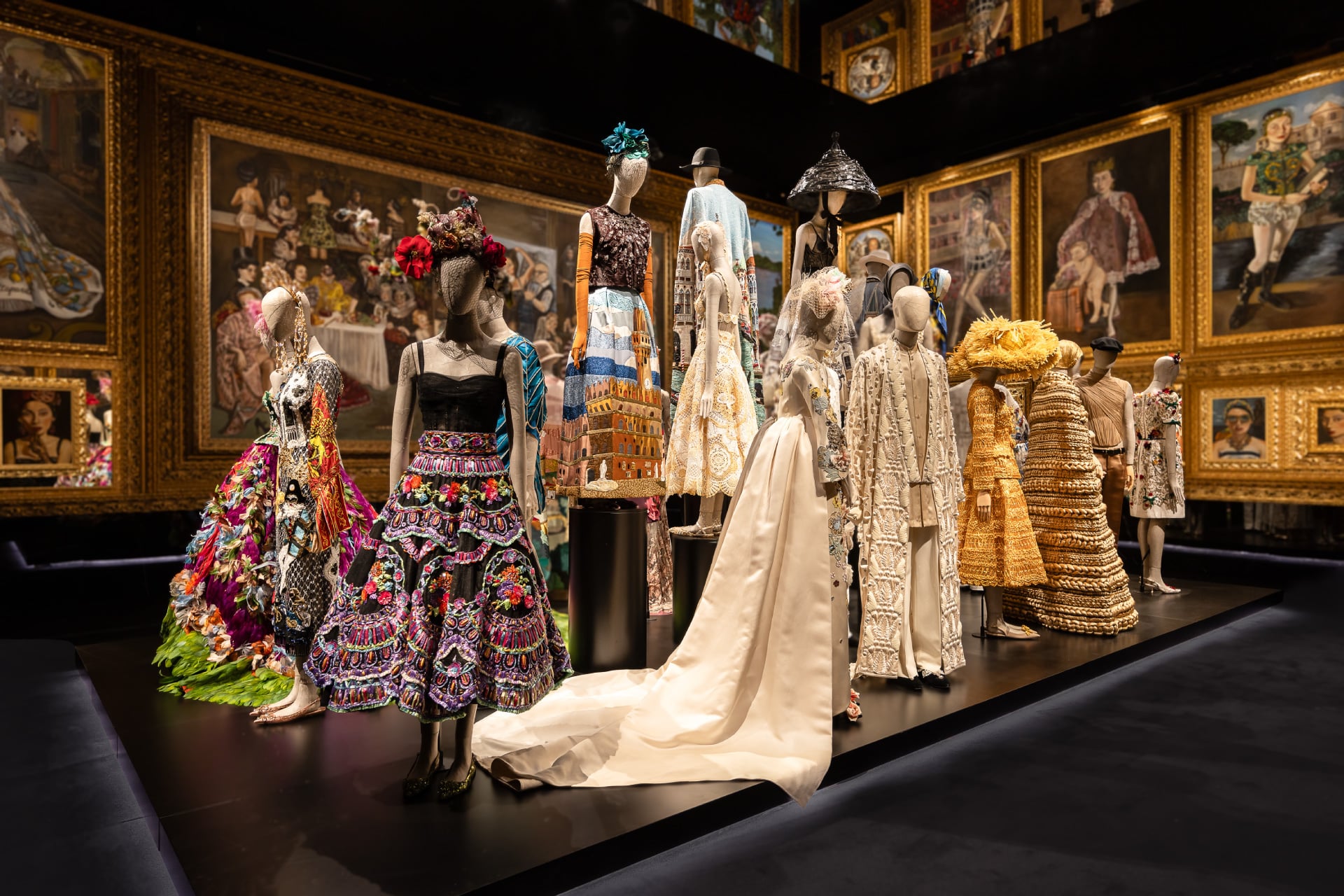 Mannequins display ornate, colorful fashion designs with floral and geometric patterns. They're set against a backdrop of framed classical paintings.