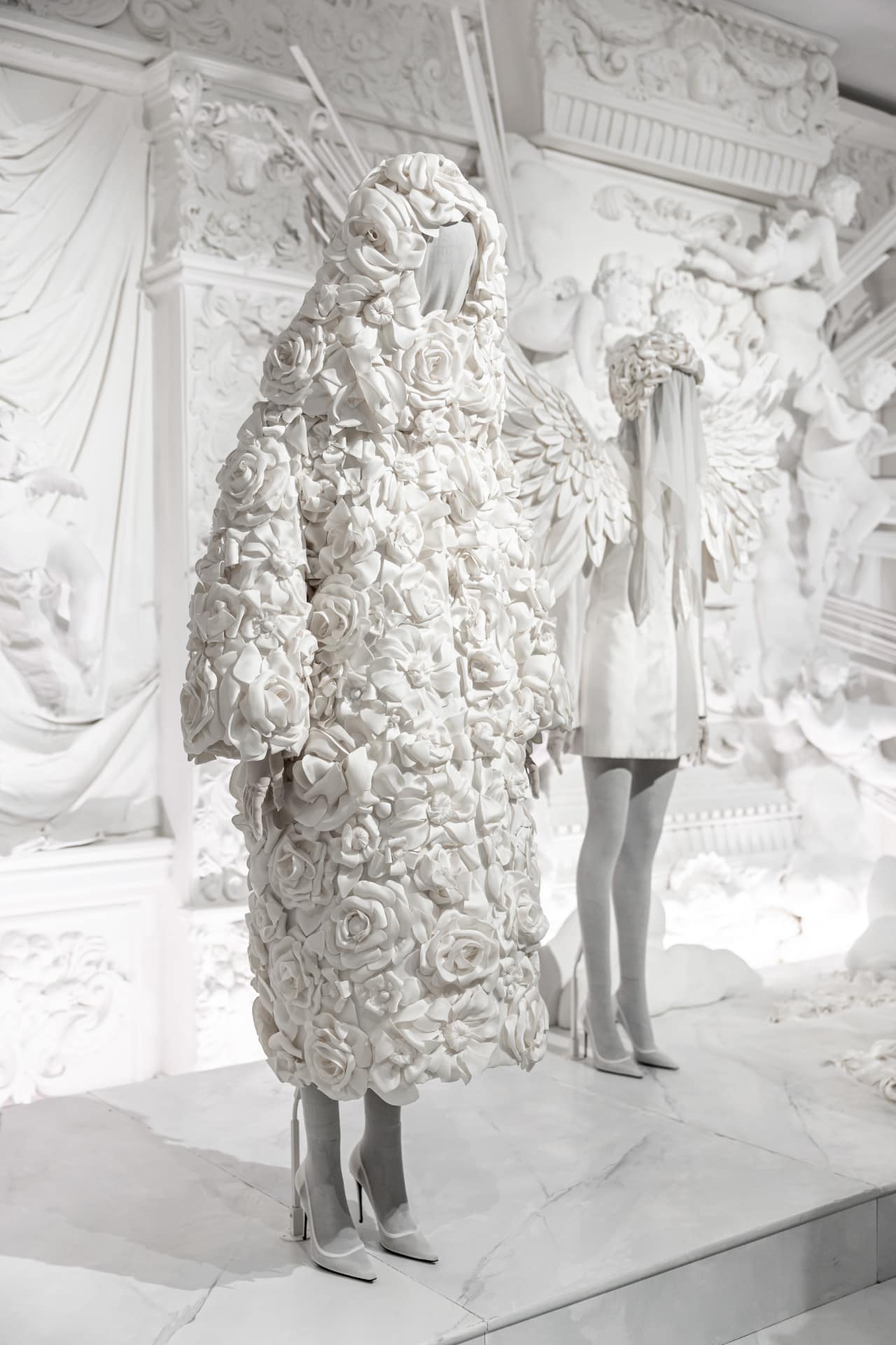 Avant-garde fashion display featuring a mannequin in a white, floral-textured coat with matching white high heels, set against a baroque-style backdrop.