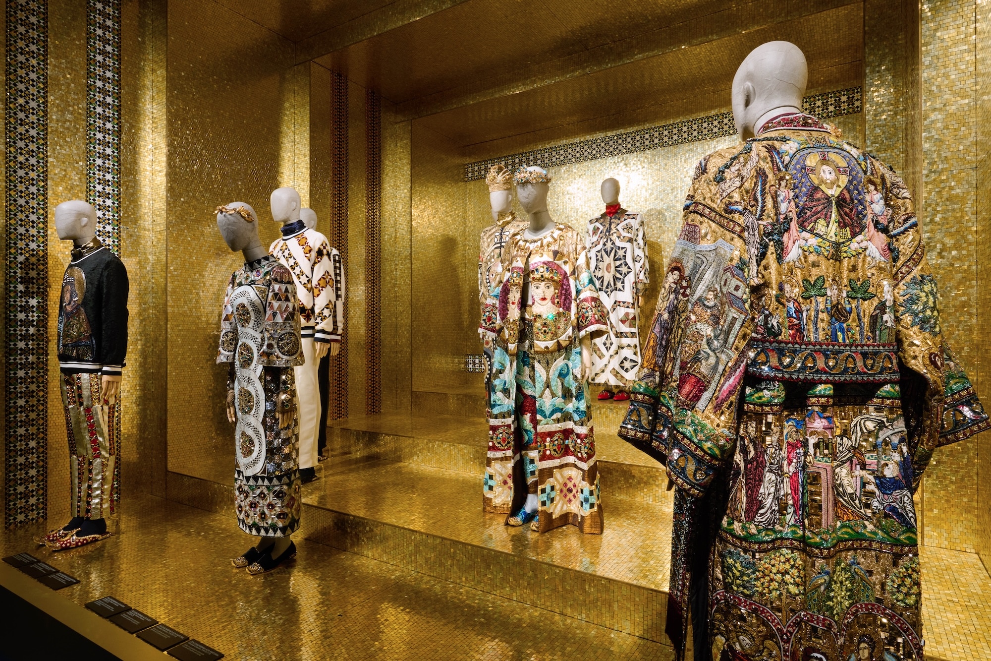 Mannequins display ornately embroidered garments in a gold-tiled space. The clothing features vibrant, intricate religious and geometric patterns.
